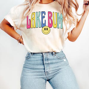 May include: Cream-colored t-shirt with the text "LAKE BUM" in colorful bubble letters and a yellow smiley face. The shirt is paired with light blue denim jeans. The overall style is casual and relaxed.