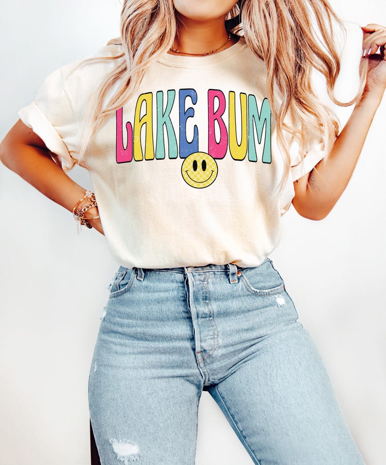 May include: Cream-colored t-shirt with the text "LAKE BUM" in colorful bubble letters and a yellow smiley face. The shirt is paired with light blue denim jeans. The overall style is casual and relaxed.