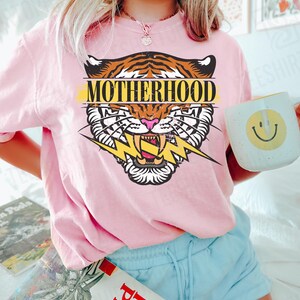 May include: Pink t-shirt with a tiger graphic and the word "MOTHERHOOD". The tiger design features orange and brown stripes, a pink nose, and a yellow lightning bolt. A white mug with a yellow smiley face is held in the hand.