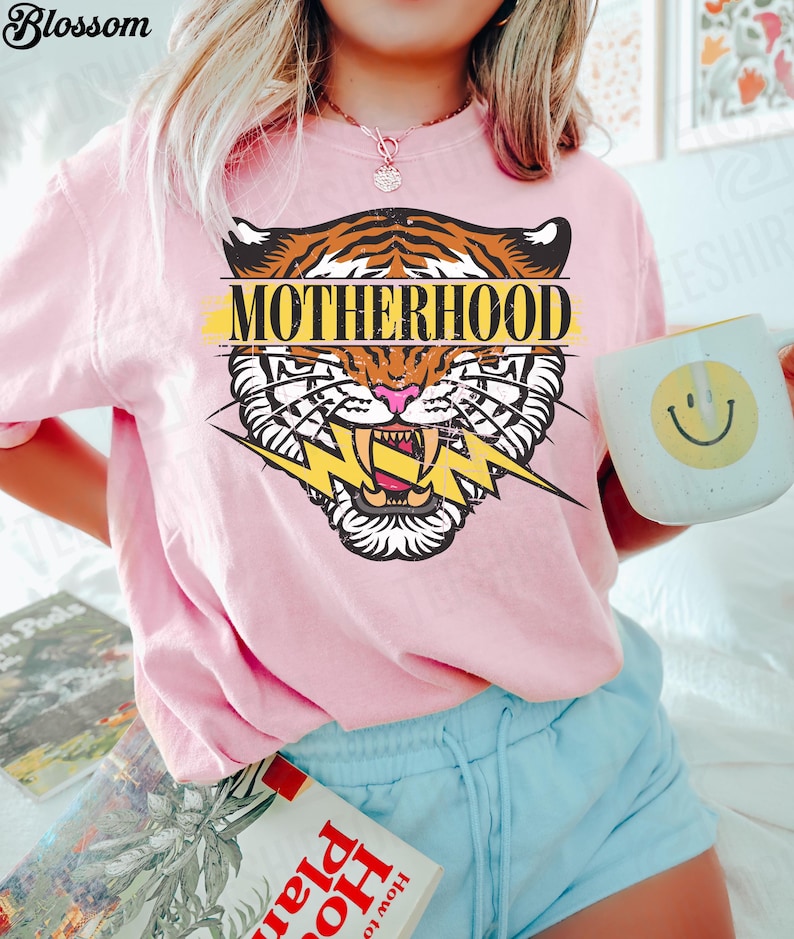 May include: Pink t-shirt with a tiger graphic and the word "MOTHERHOOD". The tiger design features orange and brown stripes, a pink nose, and a yellow lightning bolt. A white mug with a yellow smiley face is held in the hand.