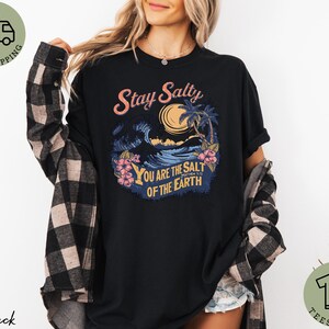 May include: Black t-shirt with a tropical graphic featuring a wave, palm trees, and the text "Stay Salty. You are the salt of the earth." The shirt has short sleeves and a relaxed fit.