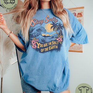 May include: A light blue t-shirt with a graphic design featuring a wave, palm trees, and the text "Stay Salty" and "You Are The Salt Of The Earth". The shirt has short sleeves and a relaxed fit.