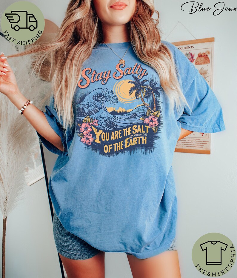 May include: A light blue t-shirt with a graphic design featuring a wave, palm trees, and the text "Stay Salty" and "You Are The Salt Of The Earth". The shirt has short sleeves and a relaxed fit.