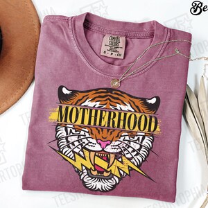 May include: A berry-colored t-shirt with a tiger graphic and the word "MOTHERHOOD" in yellow. The tiger has orange and black stripes, white teeth, and a pink nose. A gold necklace is draped across the shirt.