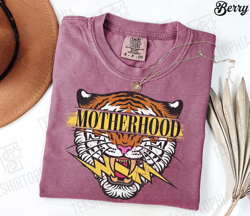 May include: A berry-colored t-shirt with a tiger graphic and the word "MOTHERHOOD" in yellow. The tiger has orange and black stripes, white teeth, and a pink nose. A gold necklace is draped across the shirt.