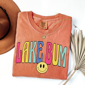 May include: Coral-colored t-shirt with the text "LAKE BUM" in colorful, retro-style lettering. A yellow smiley face with a checkered pattern is below the text. A gold necklace and a brown hat are also in the image.