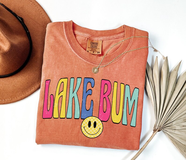 May include: Coral-colored t-shirt with the text "LAKE BUM" in colorful, retro-style lettering. A yellow smiley face with a checkered pattern is below the text. A gold necklace and a brown hat are also in the image.