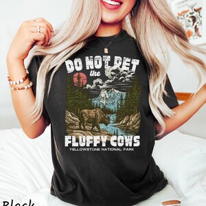 Puede incluir: Camiseta negra con un dise&ntilde;o gr&aacute;fico que presenta un bisonte, monta&ntilde;as y el texto "DO NOT PET the FLUFFY COWS Yellowstone National Park". El dise&ntilde;o incluye un sol rojo y un r&iacute;o.