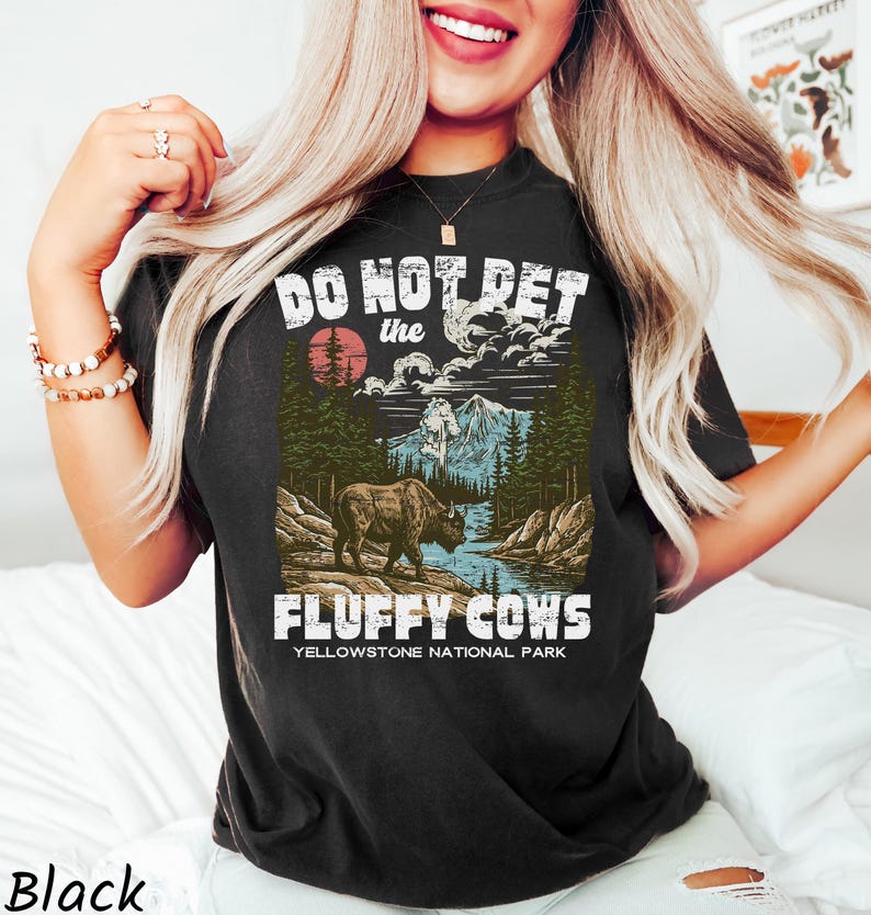 Puede incluir: Camiseta negra con un dise&ntilde;o gr&aacute;fico que presenta un bisonte, monta&ntilde;as y el texto "DO NOT PET the FLUFFY COWS Yellowstone National Park". El dise&ntilde;o incluye un sol rojo y un r&iacute;o.
