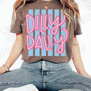 K&ouml;nnte beinhalten: Braunes T-Shirt mit dem Schriftzug "Dilly Daly" in Rosa, wei&szlig; umrandet, &uuml;ber blauen vertikalen Streifen. Das Shirt wird mit blauen Jeans und wei&szlig;en Turnschuhen getragen. Das Wort "Espresso" befindet sich in der oberen rechten Ecke.
