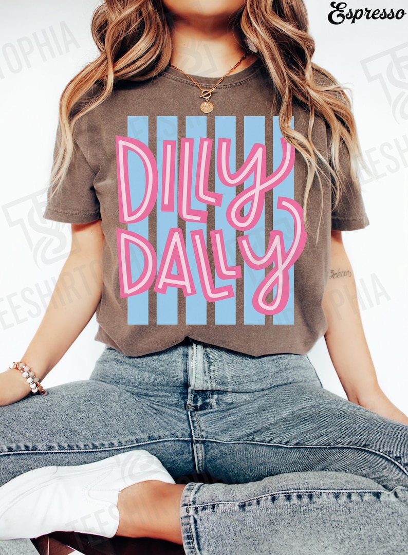 K&ouml;nnte beinhalten: Braunes T-Shirt mit dem Schriftzug "Dilly Daly" in Rosa, wei&szlig; umrandet, &uuml;ber blauen vertikalen Streifen. Das Shirt wird mit blauen Jeans und wei&szlig;en Turnschuhen getragen. Das Wort "Espresso" befindet sich in der oberen rechten Ecke.