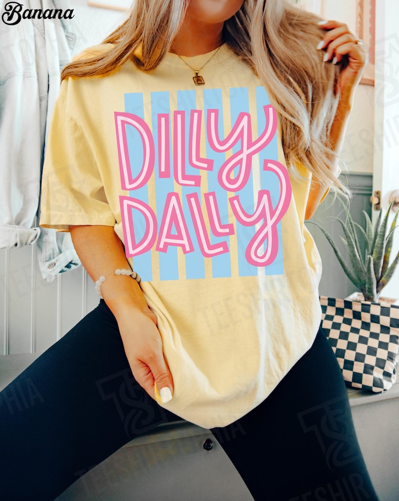 K&ouml;nnte beinhalten: Hellgelbes T-Shirt mit dem Text "DILLY DALLY" in Rosa und Blau. Der Text ist mittig auf dem Shirt platziert, mit einem hellblauen Streifenhintergrund. Das Shirt hat einen l&auml;ssigen Stil.