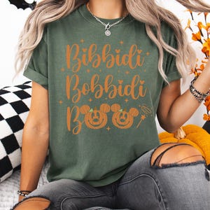 May include: An olive green t-shirt with the words "Bibbidi Bobbidi Boo" in orange script. Below the text are two jack-o'-lanterns with Mickey Mouse ears and a magic wand. The person is wearing gray jeans.