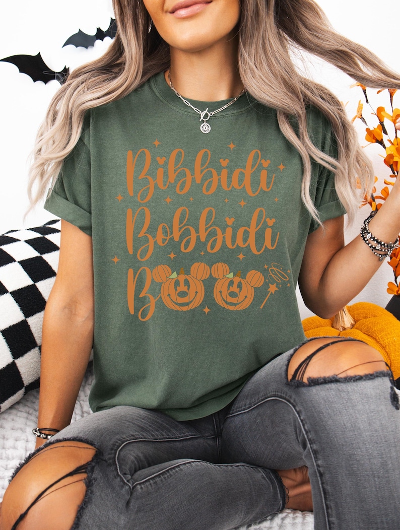 May include: An olive green t-shirt with the words "Bibbidi Bobbidi Boo" in orange script. Below the text are two jack-o'-lanterns with Mickey Mouse ears and a magic wand. The person is wearing gray jeans.