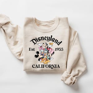 K&ouml;nnte beinhalten: Cremefarbenes Sweatshirt mit einer Grafik von Disney-Figuren, darunter Micky Maus, Minnie Maus, Donald Duck und Goofy. Der Text "Disneyland Est 1955 CALIFORNIA" ist in Schwarz &uuml;ber den Figuren aufgedruckt. Das Sweatshirt hat lange &Auml;rmel.