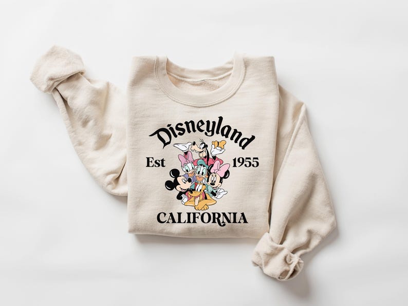 K&ouml;nnte beinhalten: Cremefarbenes Sweatshirt mit einer Grafik von Disney-Figuren, darunter Micky Maus, Minnie Maus, Donald Duck und Goofy. Der Text "Disneyland Est 1955 CALIFORNIA" ist in Schwarz &uuml;ber den Figuren aufgedruckt. Das Sweatshirt hat lange &Auml;rmel.