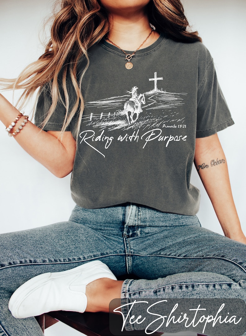 May include: A gray t-shirt with a white graphic of a person riding a horse towards a cross. The text on the shirt reads 'Proverbs 19:21' and 'Riding with Purpose'. The shirt is being worn by a person wearing blue jeans and white shoes.