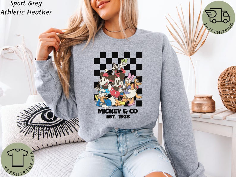 May include: Sport grey athletic heather sweatshirt featuring a colorful graphic of Mickey Mouse and friends. The design includes Mickey, Minnie, Donald Duck, Daisy Duck, Goofy, and Pluto, with the text "Mickey & Co. EST. 1928".