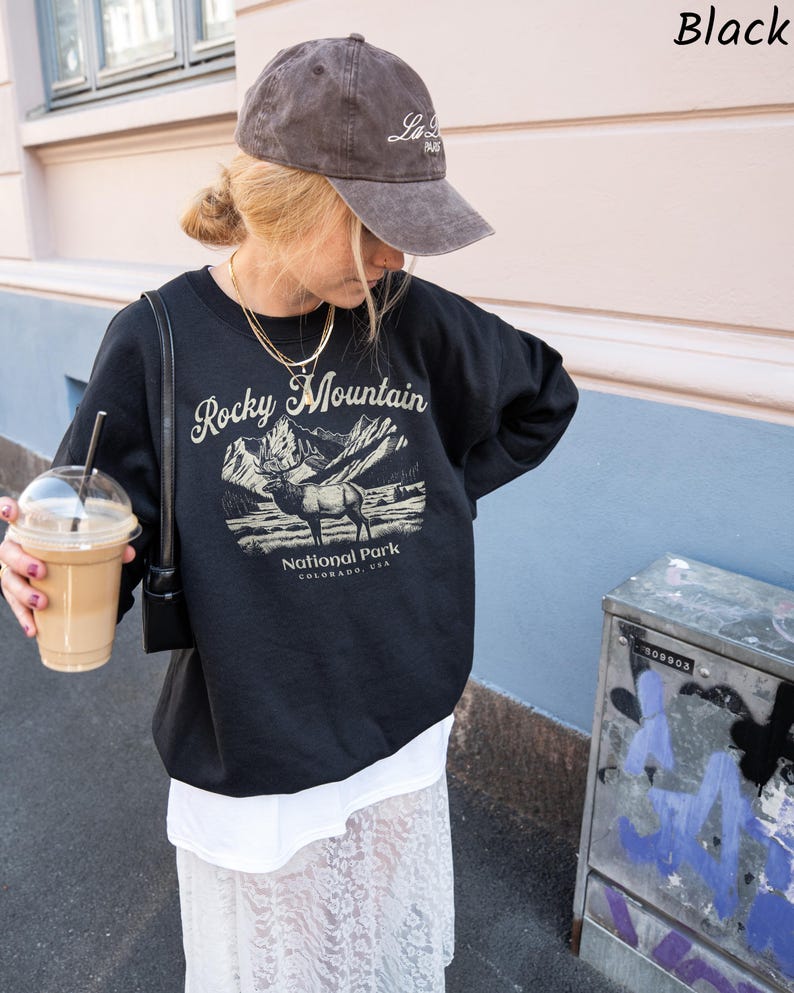 May include: A white ceramic mug with a black handle and the text "But First, Coffee" in a bold, black font. The mug is set against a blurred background, suggesting a cozy setting.