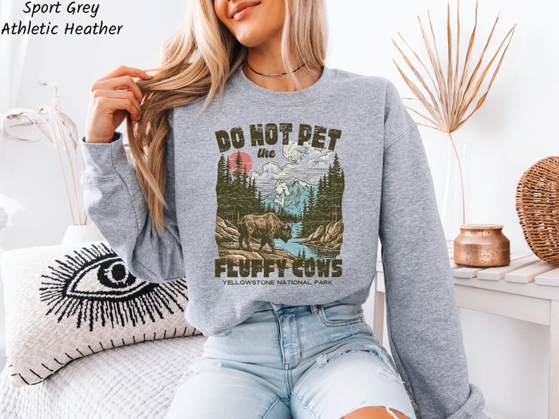 Puede incluir: Sudadera deportiva gris jaspeada con un gr&aacute;fico que dice "Do Not Pet the Fluffy Cows" sobre un paisaje del Parque Nacional de Yellowstone. El dise&ntilde;o presenta un bisonte, &aacute;rboles, monta&ntilde;as y un r&iacute;o. La sudadera es de cuello redondo y manga larga.