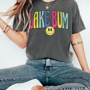 May include: Dark gray t-shirt with the text "LAKE BUM" in colorful, retro-style lettering and a yellow smiley face. The shirt is paired with blue jeans and white sneakers, creating a casual, relaxed look.