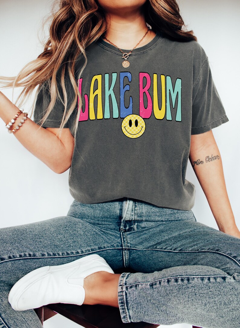 May include: Dark gray t-shirt with the text "LAKE BUM" in colorful, retro-style lettering and a yellow smiley face. The shirt is paired with blue jeans and white sneakers, creating a casual, relaxed look.