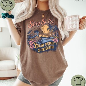 May include: Brown t-shirt with a graphic design featuring the text "Stay Salty" above a beach scene with waves, a sun, and palm trees. The text "You are the salt of the earth" is below the beach scene. The shirt has a relaxed fit.