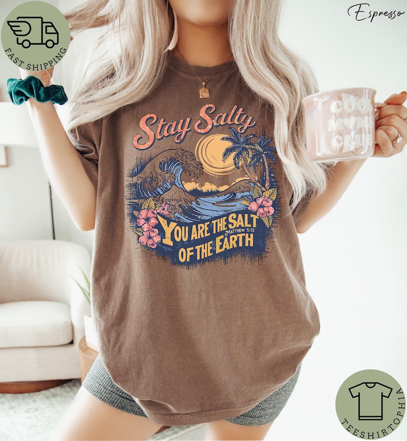 May include: Brown t-shirt with a graphic design featuring the text "Stay Salty" above a beach scene with waves, a sun, and palm trees. The text "You are the salt of the earth" is below the beach scene. The shirt has a relaxed fit.