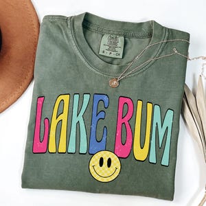 May include: An olive green t-shirt with the words "LAKE BUM" in colorful, retro-style lettering. A yellow smiley face with a checkered pattern is below the text. A gold necklace is draped across the shirt.