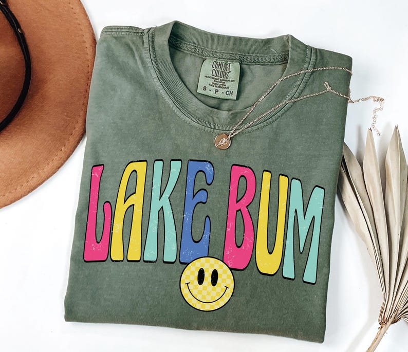 May include: An olive green t-shirt with the words "LAKE BUM" in colorful, retro-style lettering. A yellow smiley face with a checkered pattern is below the text. A gold necklace is draped across the shirt.
