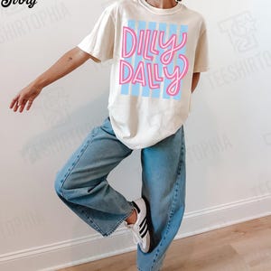 K&ouml;nnte beinhalten: Cremefarbenes T-Shirt mit dem Schriftzug "Dilly Dally" in rosa Schrift &uuml;ber hellblauen vertikalen Streifen. Die Person tr&auml;gt hellblaue Jeans und wei&szlig;e Turnschuhe mit schwarzen Streifen.