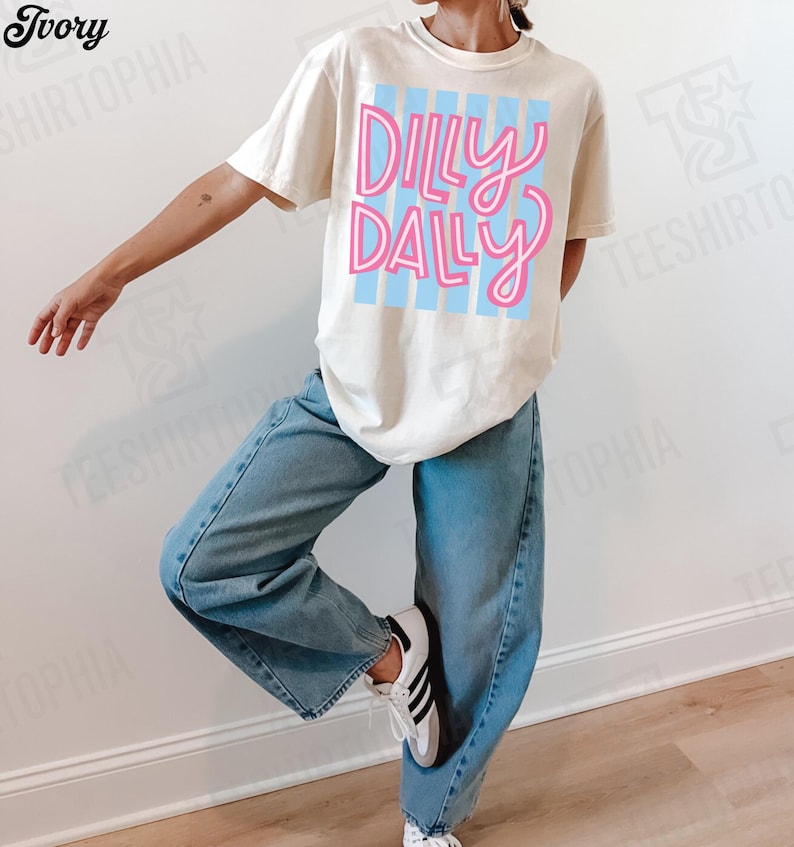 K&ouml;nnte beinhalten: Cremefarbenes T-Shirt mit dem Schriftzug "Dilly Dally" in rosa Schrift &uuml;ber hellblauen vertikalen Streifen. Die Person tr&auml;gt hellblaue Jeans und wei&szlig;e Turnschuhe mit schwarzen Streifen.