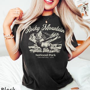 May include: Black t-shirt featuring a graphic of a Rocky Mountain National Park elk with mountains in the background. The text "Rocky Mountain National Park Colorado, USA" is printed on the shirt. The shirt is a dark color.