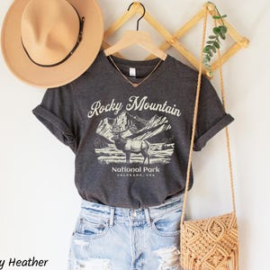 May include: Dark grey heather t-shirt with a graphic of the Rocky Mountains and an elk. The shirt is paired with light blue denim shorts, a tan hat, and a woven crossbody bag. The text on the shirt reads "Rocky Mountain National Park, Colorado, USA."