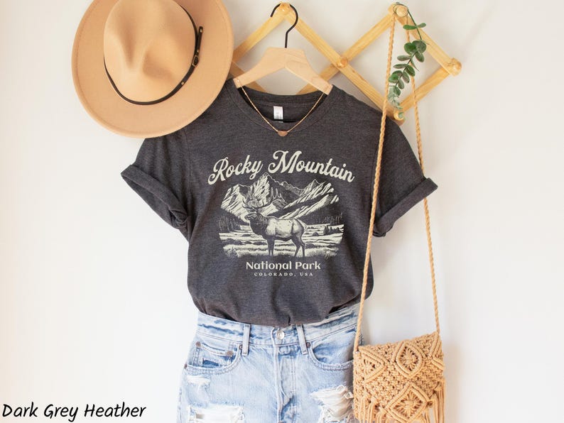 May include: Dark grey heather t-shirt with a graphic of the Rocky Mountains and an elk. The shirt is paired with light blue denim shorts, a tan hat, and a woven crossbody bag. The text on the shirt reads "Rocky Mountain National Park, Colorado, USA."