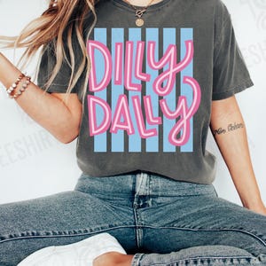 K&ouml;nnte beinhalten: Dunkelgraues T-Shirt mit dem Schriftzug "Dilly Dally" in rosa Schrift &uuml;ber blauen vertikalen Streifen. Ein l&auml;ssiges Oberteil f&uuml;r den Alltag.