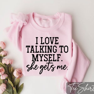 May include: A light pink sweatshirt with the text "I LOVE TALKING TO MYSELF. she gets me" in black. The sweatshirt is folded and placed on a white surface, with pink flowers in the corner.