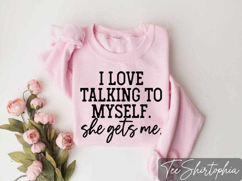 May include: A light pink sweatshirt with the text "I LOVE TALKING TO MYSELF. she gets me" in black. The sweatshirt is folded and placed on a white surface, with pink flowers in the corner.
