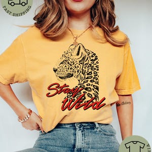 May include: Mustard-colored t-shirt featuring a leopard graphic and the words "Stay Wild" in red script. The leopard design is black and detailed. The shirt is worn with blue jeans.
