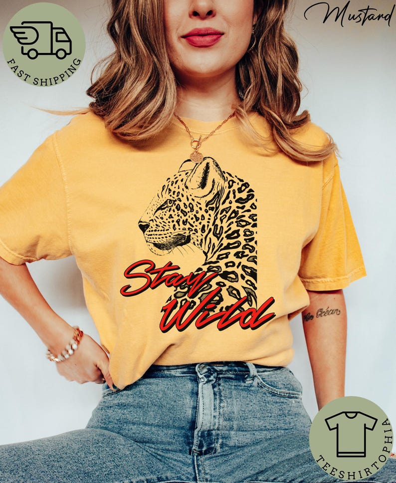 May include: Mustard-colored t-shirt featuring a leopard graphic and the words "Stay Wild" in red script. The leopard design is black and detailed. The shirt is worn with blue jeans.