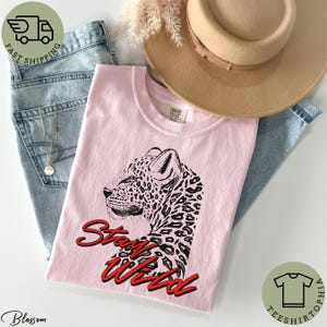 May include: A light pink t-shirt featuring a black and white leopard illustration with the words "Stay Wild" in red script. The shirt is folded with a pair of blue jeans, a tan hat, and a gold necklace.