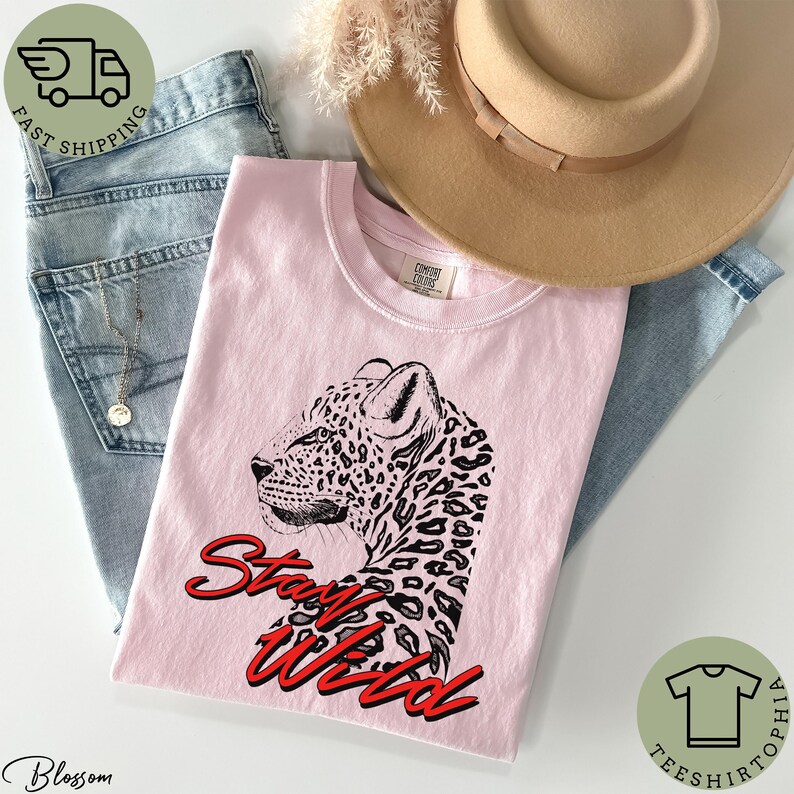May include: A light pink t-shirt featuring a black and white leopard illustration with the words "Stay Wild" in red script. The shirt is folded with a pair of blue jeans, a tan hat, and a gold necklace.