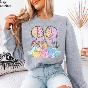 Puede incluir: Una sudadera gris claro con un gr&aacute;fico colorido de princesas de Disney y un gran lazo. Las princesas visten sus vestidos ic&oacute;nicos. El texto "Sport Grey Athletic Heather" est&aacute; en la esquina superior izquierda.
