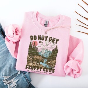 Puede incluir: Sudadera rosa claro con el texto "DO NOT PET the FLUFFY COWS" y un gr&aacute;fico de un bisonte en el Parque Nacional de Yellowstone. La sudadera est&aacute; doblada con un collar encima y colocada junto a unos vaqueros azules.
