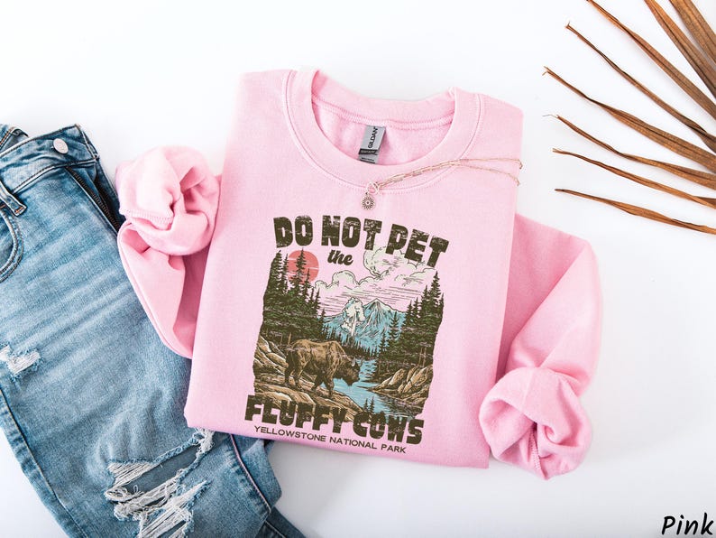 Puede incluir: Sudadera rosa claro con el texto "DO NOT PET the FLUFFY COWS" y un gr&aacute;fico de un bisonte en el Parque Nacional de Yellowstone. La sudadera est&aacute; doblada con un collar encima y colocada junto a unos vaqueros azules.