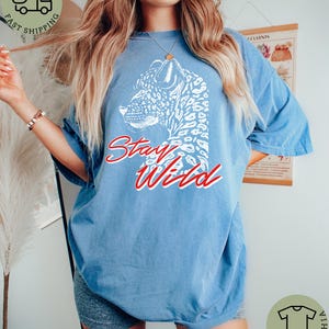 May include: A light blue t-shirt featuring a white leopard design and the words "Stay Wild" in red script. The shirt has a relaxed fit and short sleeves, perfect for casual wear.
