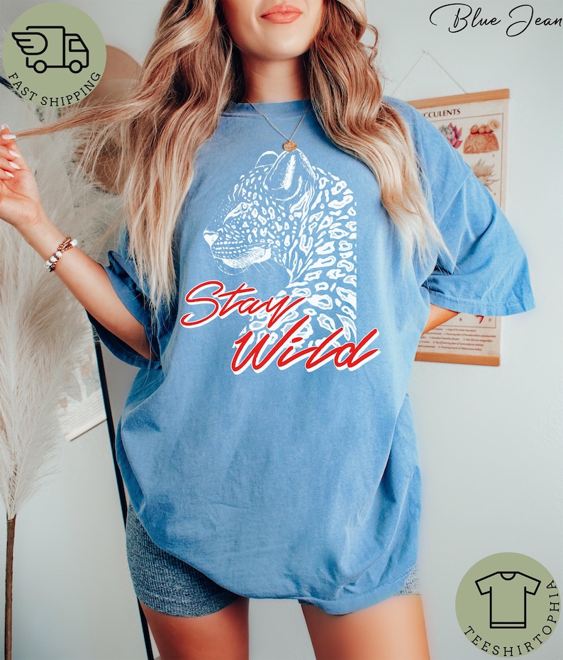 May include: A light blue t-shirt featuring a white leopard design and the words "Stay Wild" in red script. The shirt has a relaxed fit and short sleeves, perfect for casual wear.