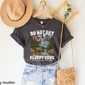 Puede incluir: Camiseta gris oscuro jaspeado con un gr&aacute;fico de un bisonte en un paisaje del Parque Nacional de Yellowstone. El texto de la camiseta dice "DO NOT PET the FLUFFY COWS". Tambi&eacute;n se ven un sombrero beige y un bolso tejido.