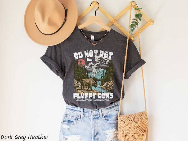 Puede incluir: Camiseta gris oscuro jaspeado con un gr&aacute;fico de un bisonte en un paisaje del Parque Nacional de Yellowstone. El texto de la camiseta dice "DO NOT PET the FLUFFY COWS". Tambi&eacute;n se ven un sombrero beige y un bolso tejido.