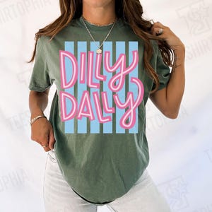 K&ouml;nnte beinhalten: Olivgr&uuml;nes T-Shirt mit dem Schriftzug "Dilly Dally" in rosa Schrift auf hellblau gestreiftem Hintergrund. Das T-Shirt hat einen l&auml;ssigen Stil und ist f&uuml;r den Alltag geeignet.